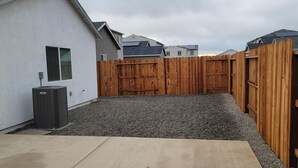 Un jardin arrière d’une maison, avec une terrasse en béton, une clôture en bois et une zone recouverte de gravier.