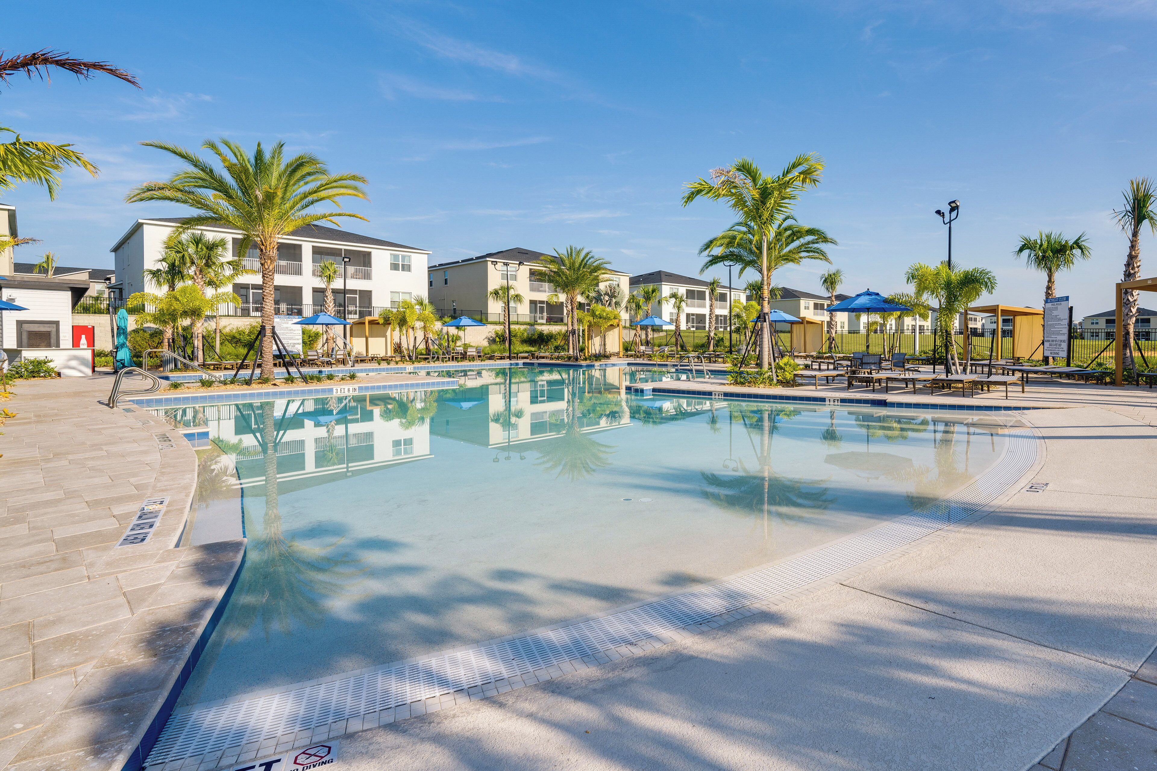Una piscina con bordo piastrellato, circondata da palme e un edificio con balconi.