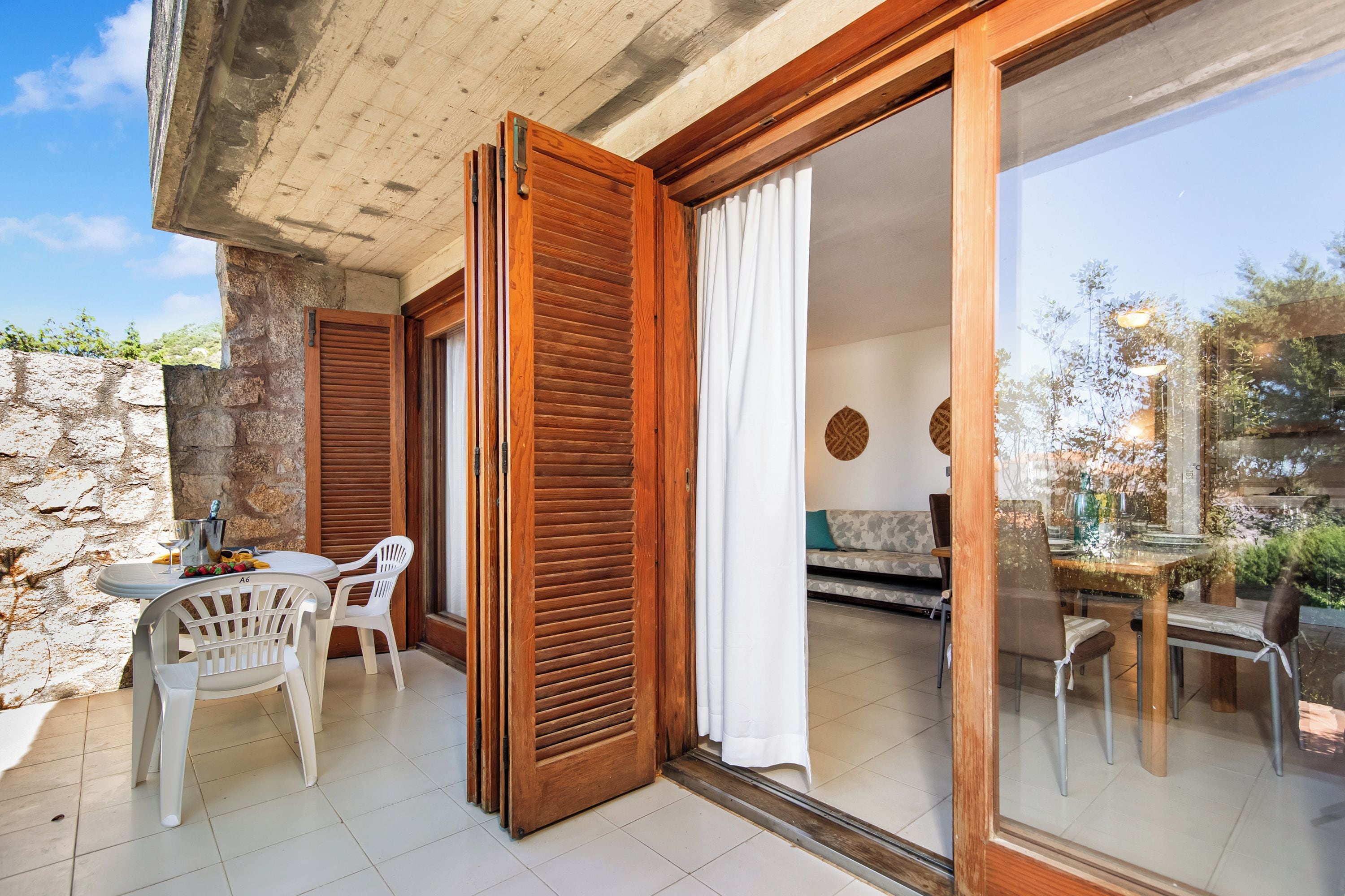 Una habitación con pared de piedra, contraventanas de madera, una mesa de comedor con sillas y vistas a través de un ventanal.