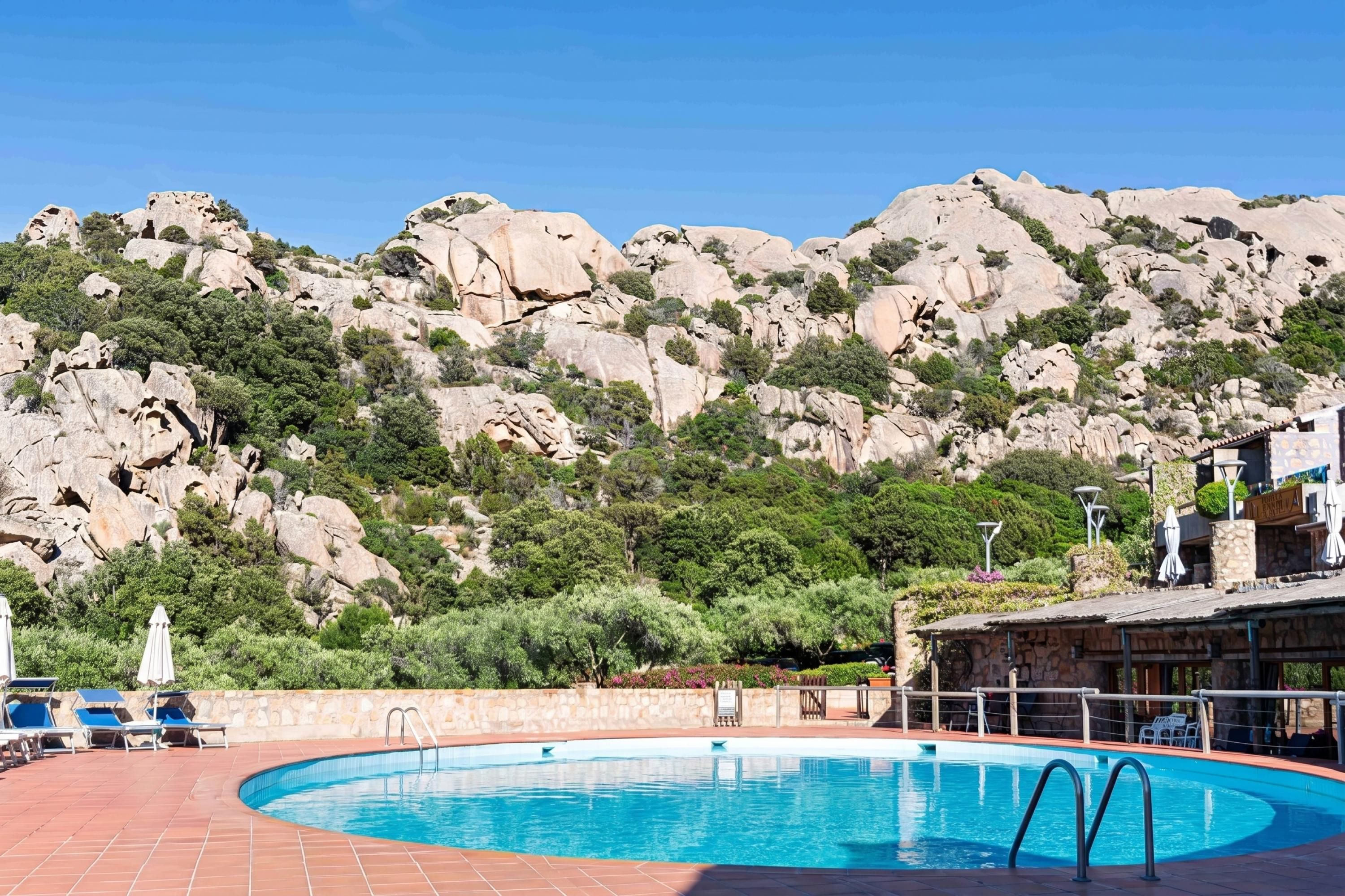 A pool with a stone wall and lounge chairs, surrounded by rocky hills and greenery.