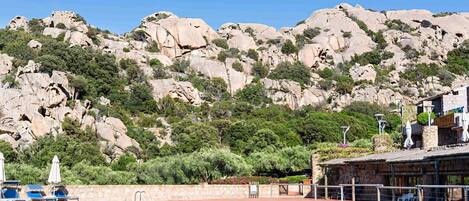 A pool with a stone wall and lounge chairs, surrounded by rocky hills and greenery.