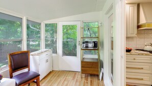 Une cuisine avec des meubles blancs, une chaise en bois, un four à micro-ondes et une vue sur la verdure à l’extérieur.