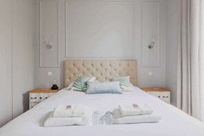 Een net opgemaakt bed met een gestoffeerd hoofdbord, twee nachtkastjes met lampen, en een stapel handdoeken op het bed.