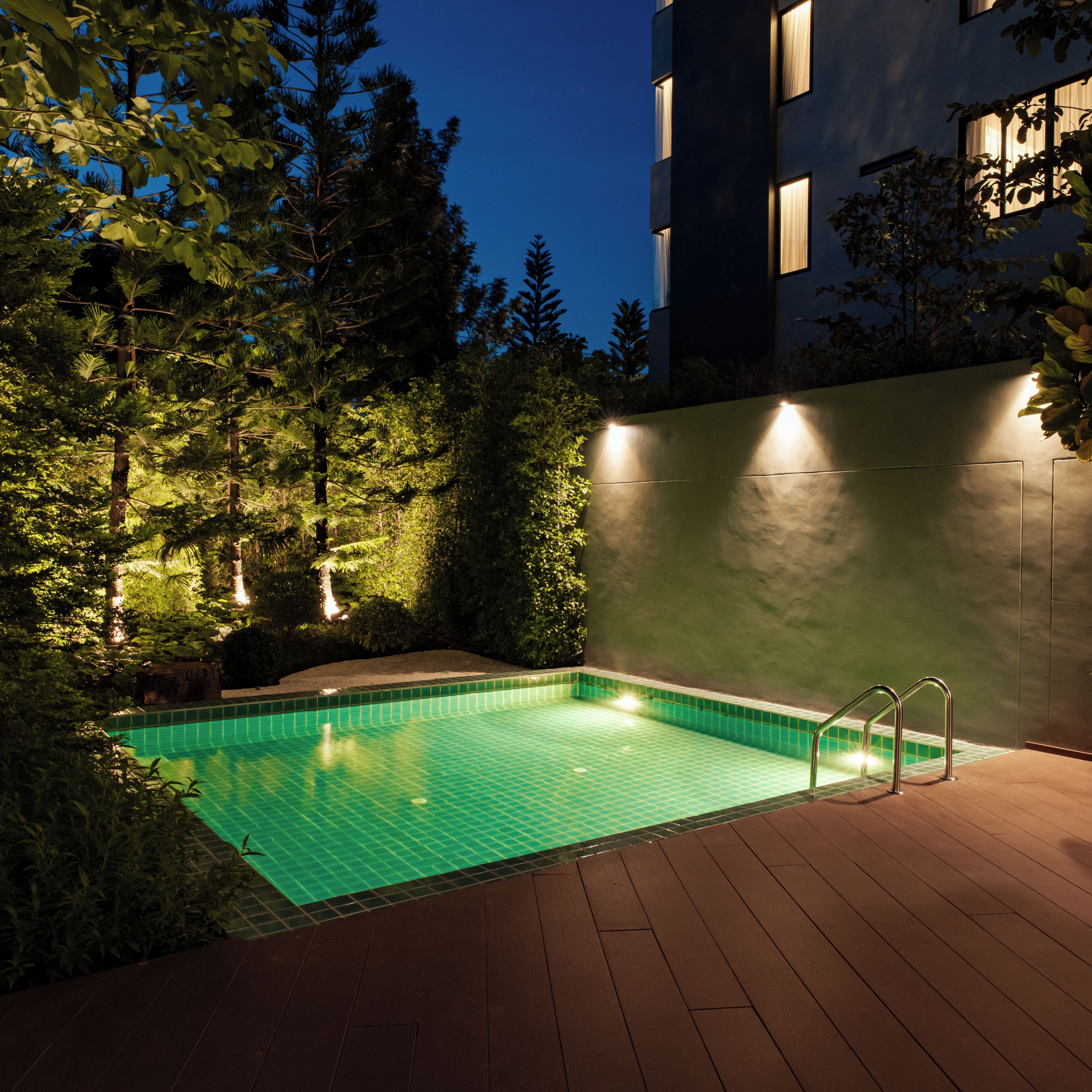 A swimming pool with a wooden deck, surrounded by trees and a building with illuminated windows.