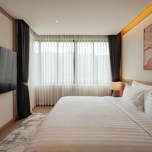 A hotel room with a large bed, a flat-screen TV, a bedside table with a lamp, and a window with sheer curtains. - Zen Essence Khao Yai (Pak Chong)