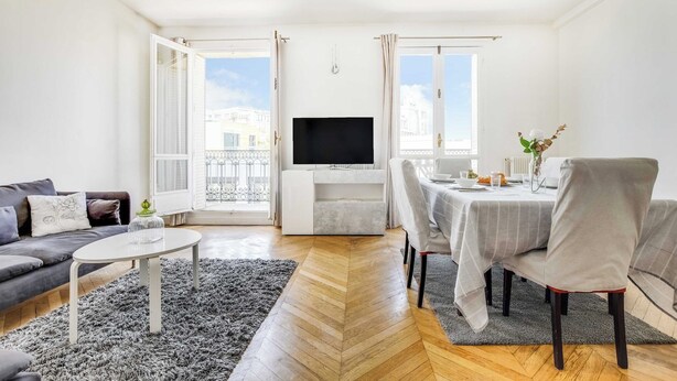 Moderni olohuone, jossa on sohva, ruokapöytä tuoleineen, taulutelevisio ja suuri ikkuna, josta on näkymä rakennuksiin.