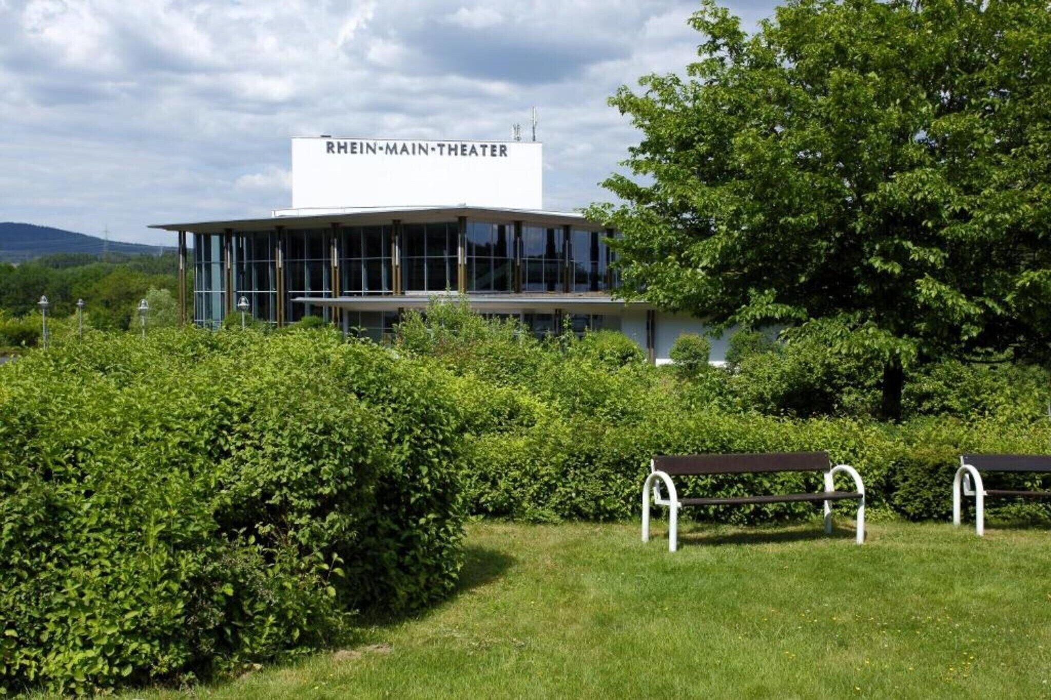 Un bâtiment moderne, portant l’inscription « RHIN-MAIN-THÉÂTRE », avec une façade en verre, entouré d’une végétation luxuriante et agrémenté d’un banc.