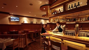 Zona de bar con mobiliario de madera, un televisor montado en la pared y estantes con botellas.