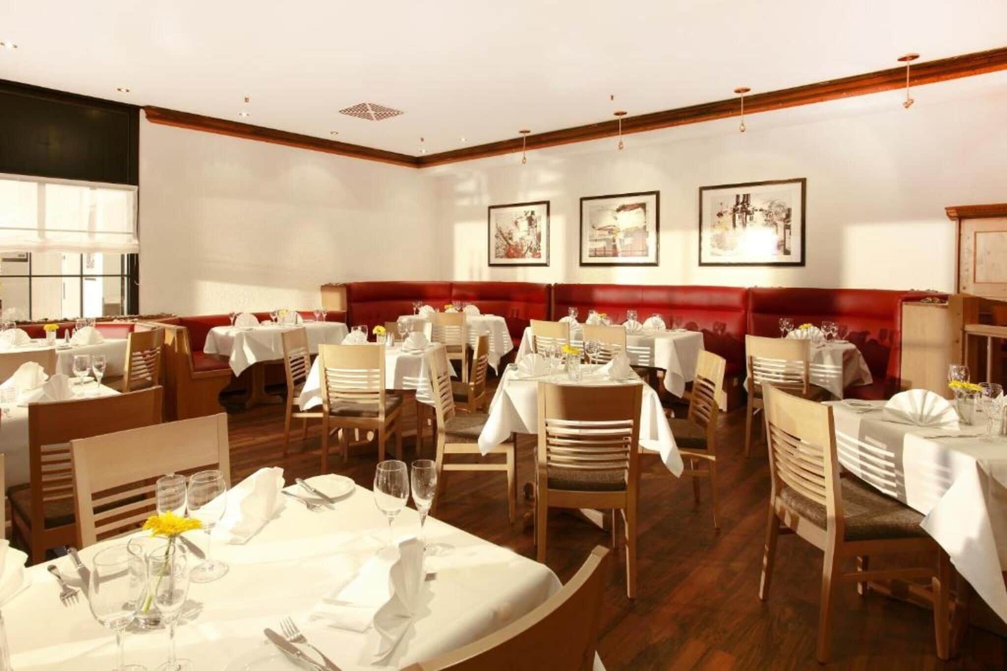 A restaurant with multiple tables set for dining, wooden chairs, and a red cushioned bench area.