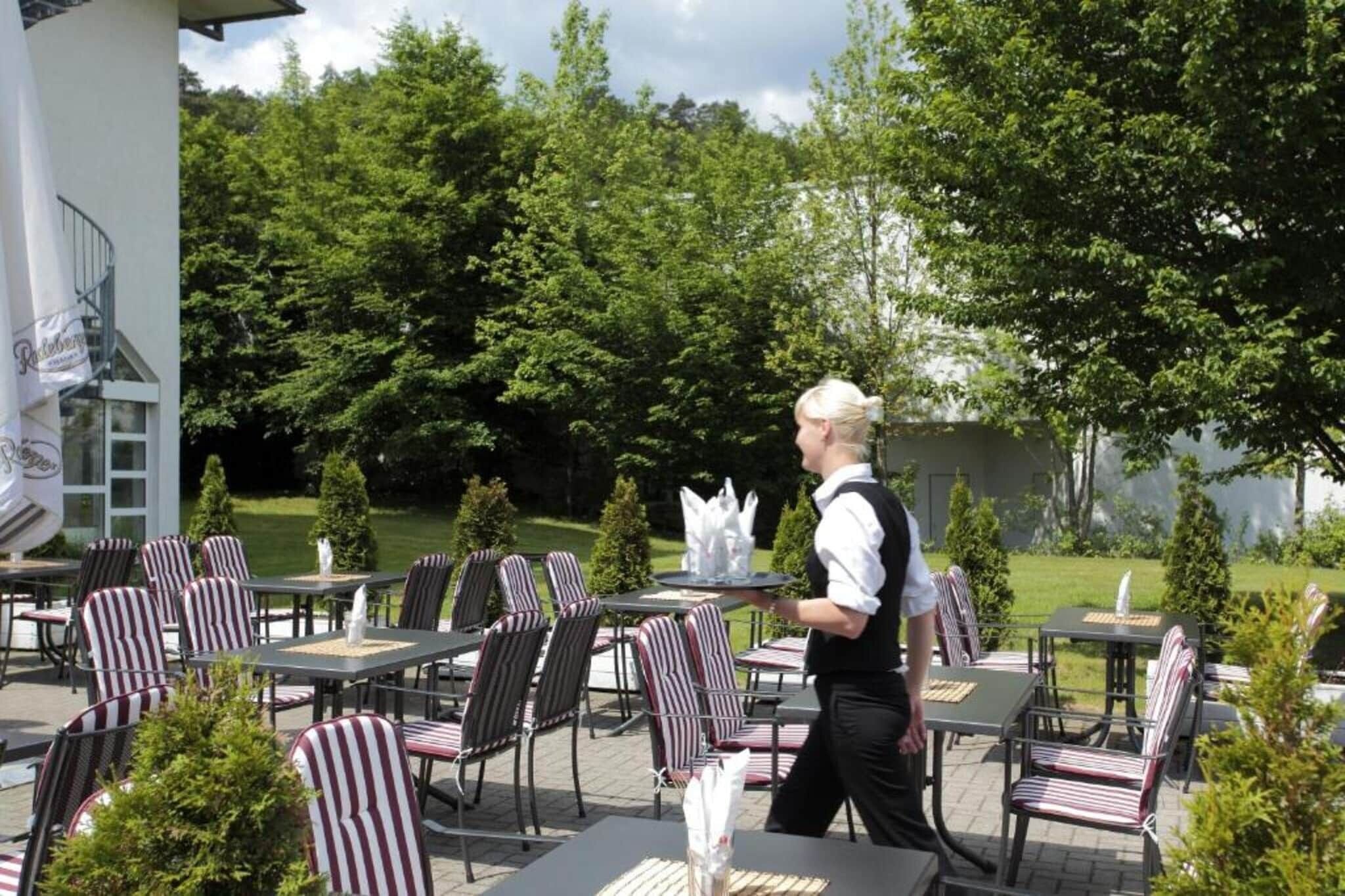 Un espace de restauration en plein air avec des chaises et des tables à rayures, un serveur qui passe, et un bâtiment à la façade blanche en arrière-plan.