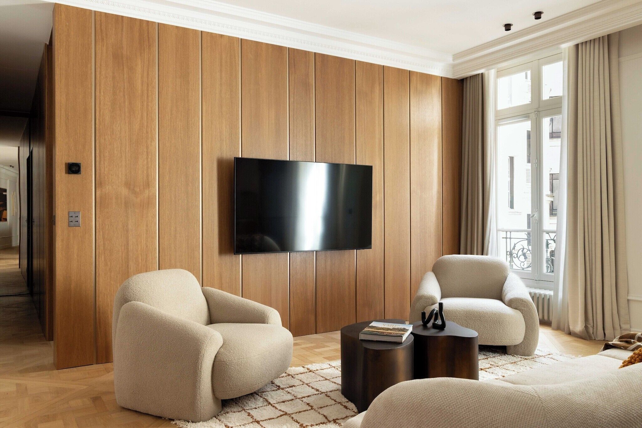 Een moderne woonkamer met een flatscreen televisie bevestigd aan een houten wandpaneel, twee beige fauteuils, een ronde salontafel en een uitzicht op het buitenlands landschap door een groot raam.