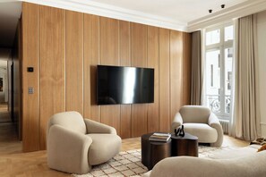 Ein modernes Wohnzimmer mit einem Flachbildfernseher an einer Wand aus Holzpaneelen, zwei beigefarbenen Sesseln, einem runden Couchtisch und Blick ins Freie durch ein großes Fenster.