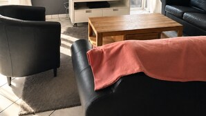 A modern living room with a flat-screen TV on a white console, black leather sofas, a wooden coffee table, and a large window with curtains. - Vacation villa with conservatory at the 't Posterbos park (Posterholt)