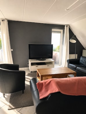 A modern living room with a flat-screen TV on a white console, black leather sofas, a wooden coffee table, and a large window with curtains. - Vacation villa with conservatory at the 't Posterbos park (Posterholt)