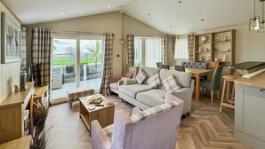 A spacious living area with a sofa, coffee table, and dining table. There is a kitchen area with a counter and cabinets. Large windows provide natural light and a view of the outdoors. - Willerby Portland - UKC9613 (Towyn)