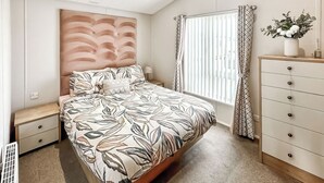 A bedroom with a bed, a dresser, a nightstand, a window with curtains, and a wall-mounted headboard. - Willerby Portland - UKC9613 (Towyn)