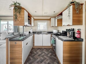 A compact kitchen with white cabinets, a herringbone-patterned floor, a built-in stove, and a window with blinds. - ABI St David - UKC9615 (Towyn)