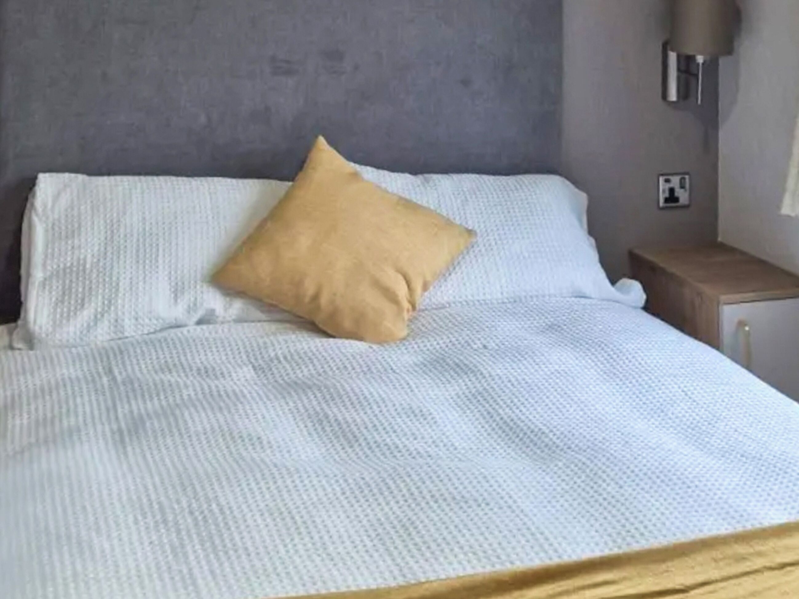 En säng med ett vitt, strukturerat överkast och en beige kudde, en grå vägg, ett litet träbord vid sängen med en lampa och ett eluttag.