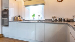 A modern kitchen with a built-in oven, a stainless steel refrigerator, a window with a plant, and a clock on the wall. - The Cottage - UKC9711 (Winster)