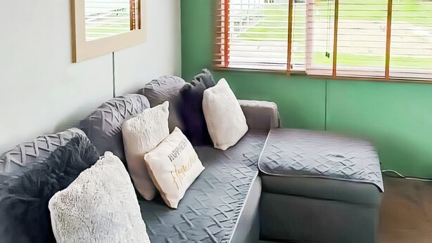 Ein modernes Wohnzimmer mit einem grauen Ecksofa, mehreren Kissen, einem gerahmten Bild an der Wand und einem Fenster mit Jalousien.