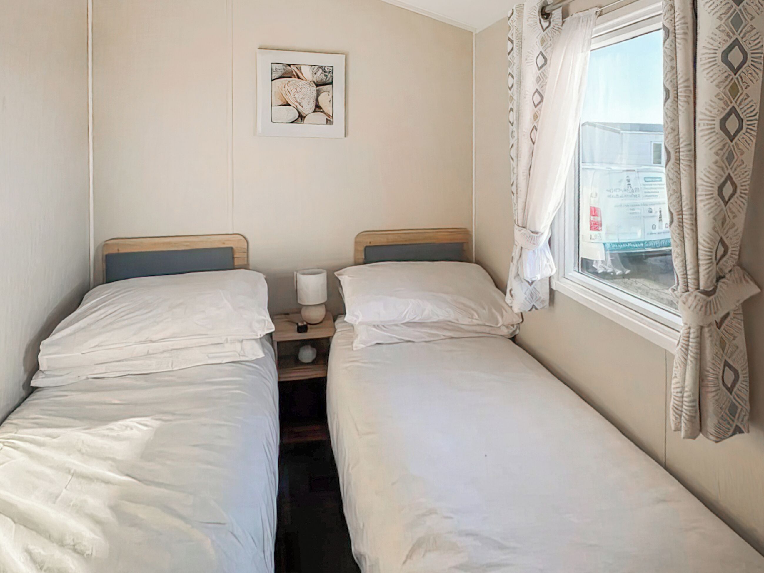 Ein Doppelzimmer mit zwei Einzelbetten, einem Nachttisch mit Lampe, einem gerahmten Bild an der Wand und einem Fenster mit Vorhängen.