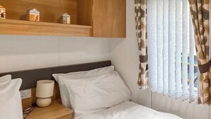 A single bed with white bedding, a wooden nightstand with a lamp, and a shelf with model houses. - Swift Escape - UKC9641 (Towyn)
