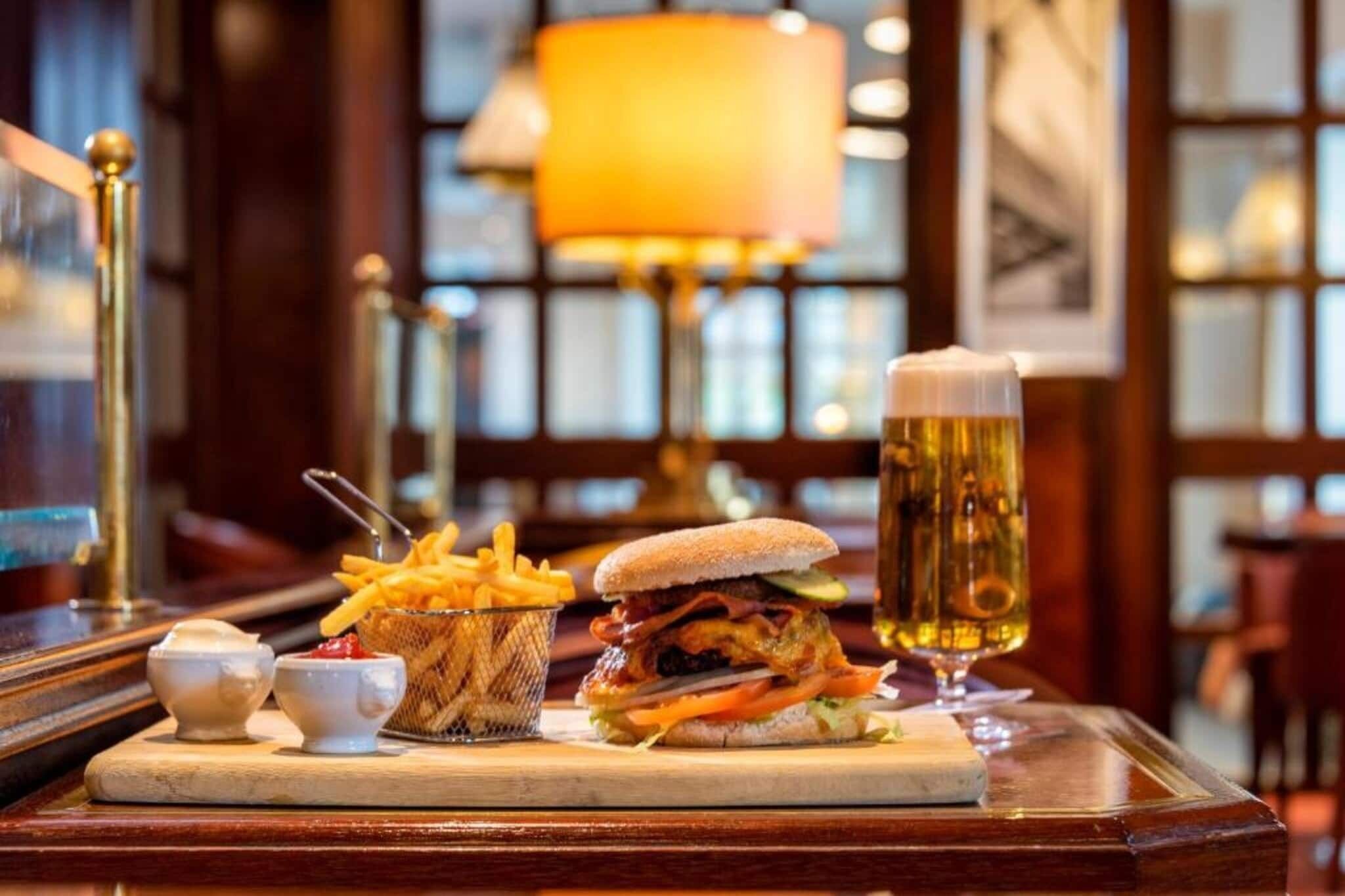 A meal with a burger, fries, and a beer glass on a wooden table in a restaurant with a warm ambiance.
