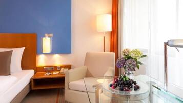 Une chambre d’hôtel avec un lit, un bureau, une chaise, une lampe et une table en verre sur laquelle se trouvent des fruits.
