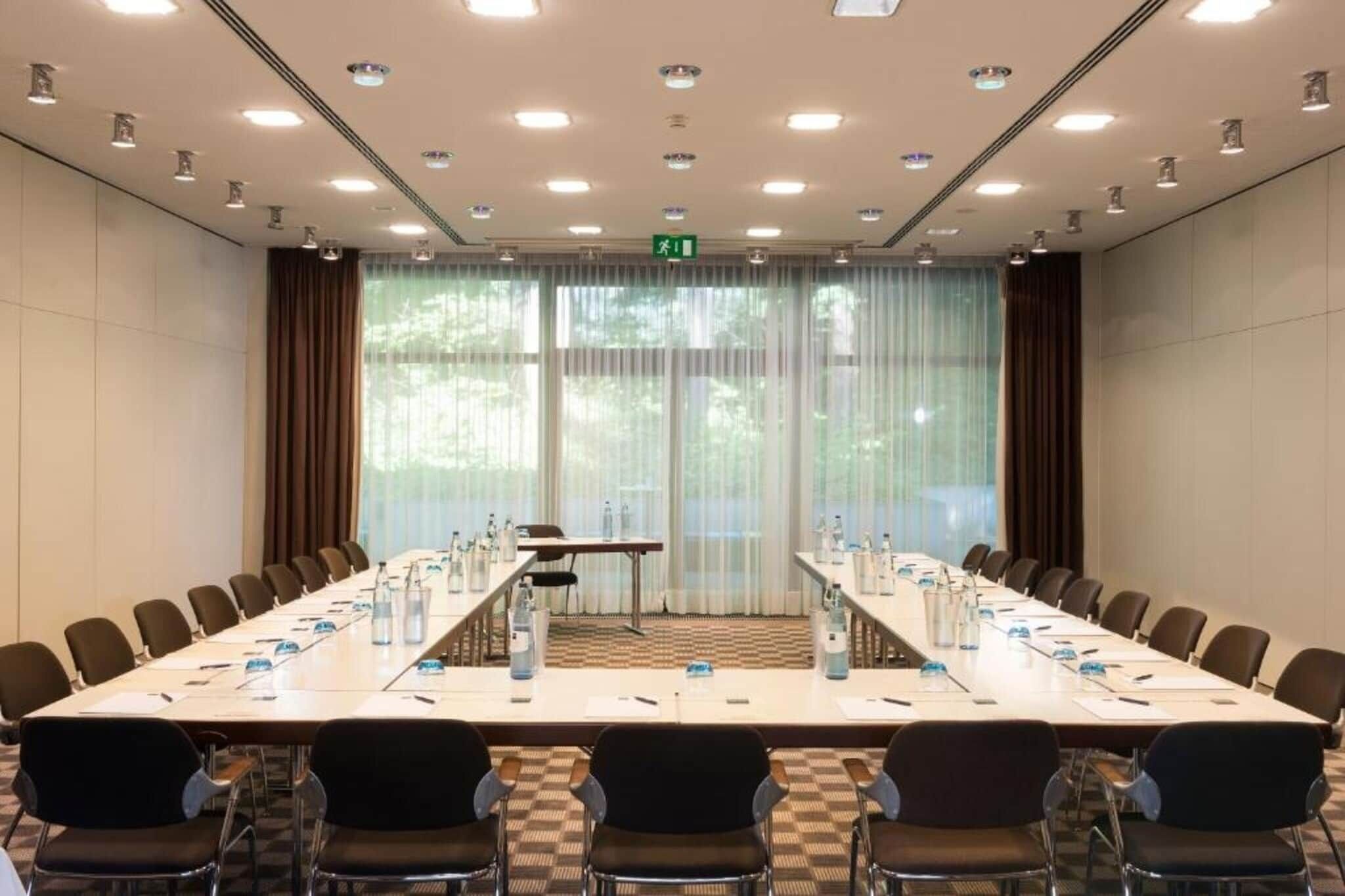 Una sala conferenze con un lungo tavolo, sedie, bottiglie d'acqua e vista su alberi attraverso ampie finestre.