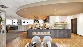 A modern hotel lobby with a central bar area, seating arrangement, and a visible kitchen in the background.