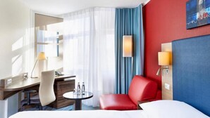 A hotel room with a bed, a desk with a chair, a small table with a lamp, and a red armchair.