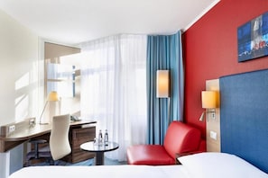 A hotel room with a bed, a desk with a chair, a small table with a lamp, and a red armchair.
