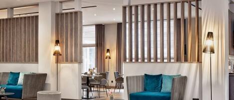 A modern hotel lobby with a seating area, a dining area, and a kitchen visible in the background.