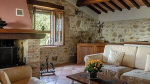 A rustic living room with a stone wall, wooden beams, a fireplace, a sofa, and a coffee table with flowers. - Villa Daluma, Private Pool in Todi (Todi)