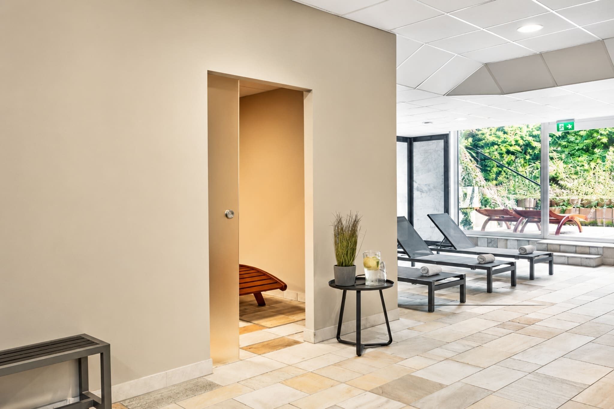 A modern indoor area with a bench, lounge chairs, and a small table with a plant and drink.