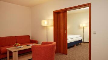 Una habitación de hotel con puerta de madera, un sofá rojo, una mesita con lámpara y una cama con colchón blanco.