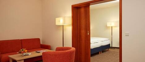 A hotel room with a wooden door, a red sofa, a small table with a lamp, and a bed with a white mattress.