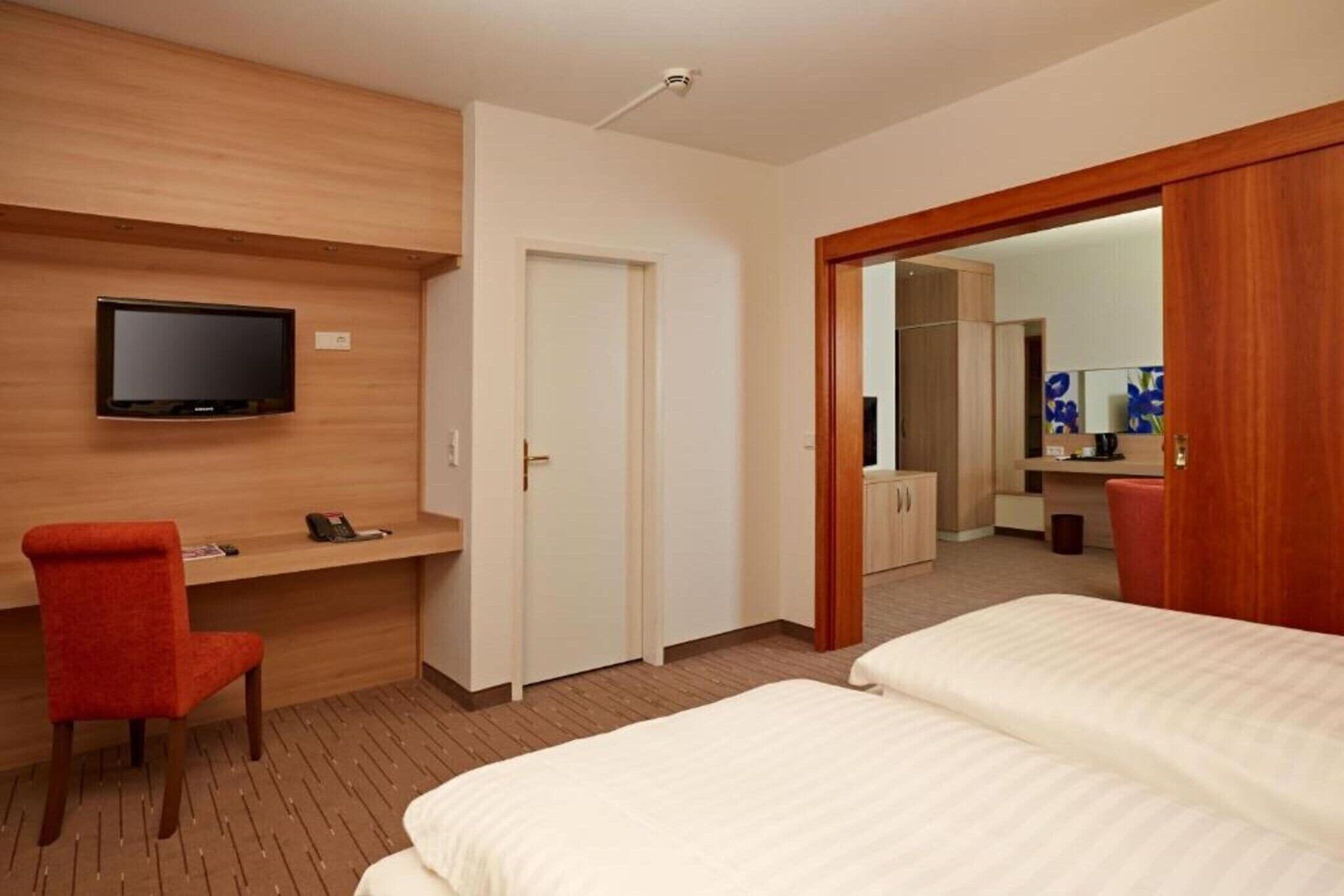 Habitación de hotel con cama, escritorio con silla, televisor montado en la pared y puerta que da a otra habitación.