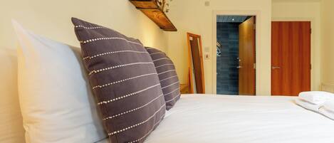 A hotel room with a bed, pillows, a wooden shelf with decorative items, and a door leading to another room.