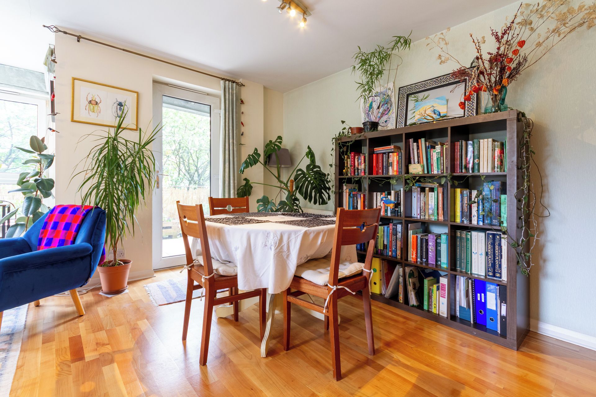 A room with a dining table, chairs, a bookshelf, and various plants.