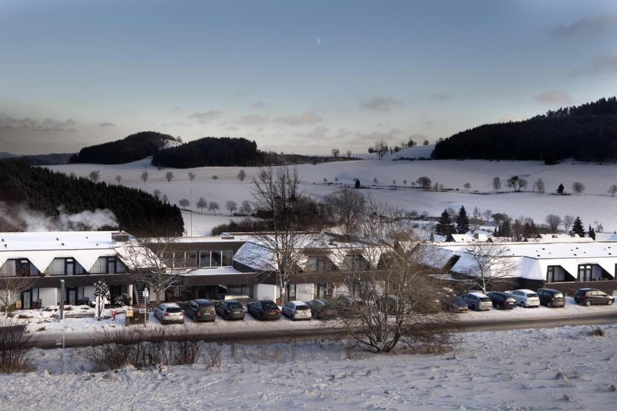A hotel with multiple buildings, a parking lot with several cars, and a snowy landscape with hills in the background.
