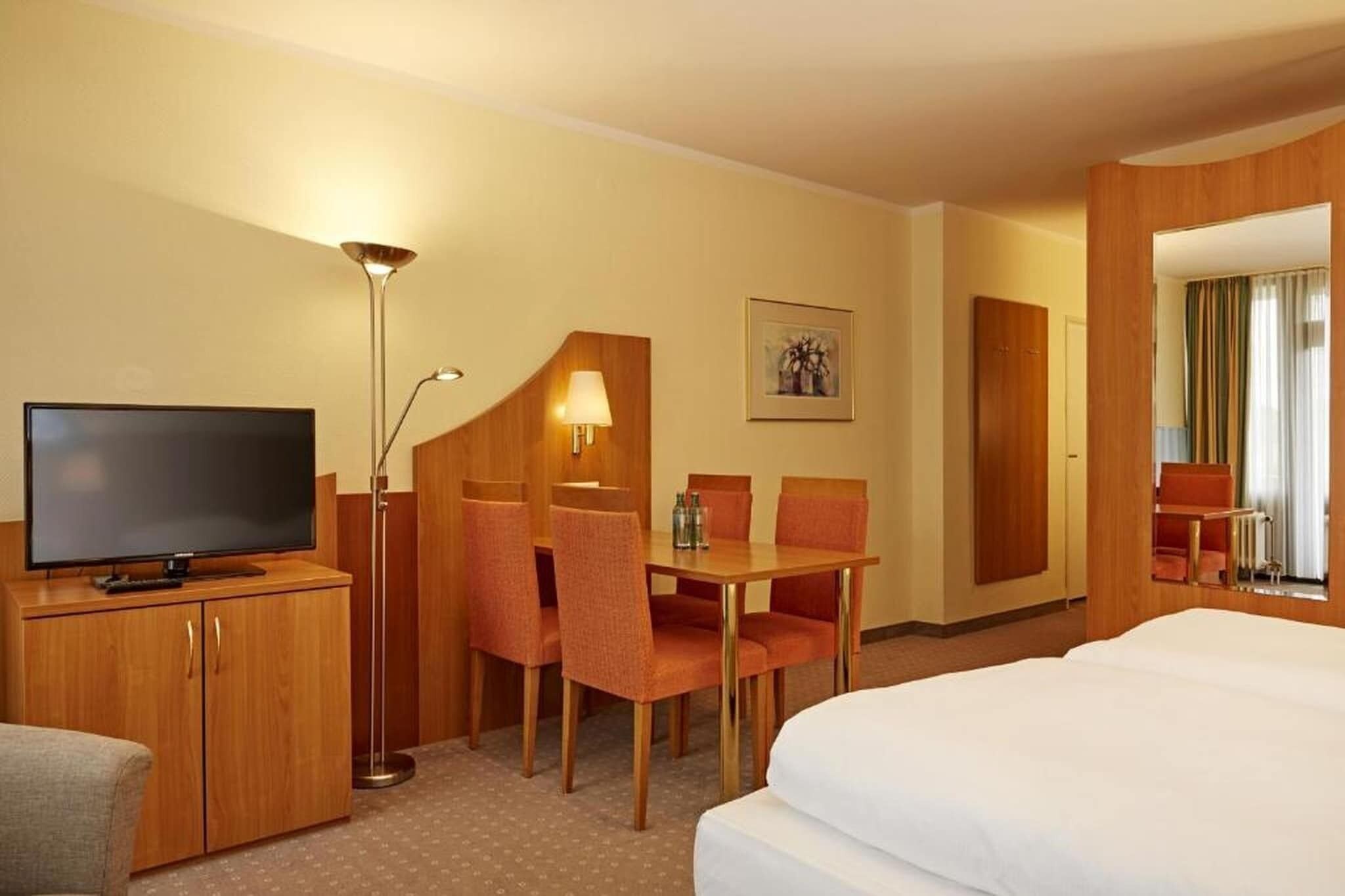 Habitación de hotel con cama, televisión, escritorio y sillas, lámpara y un cuadro en la pared.