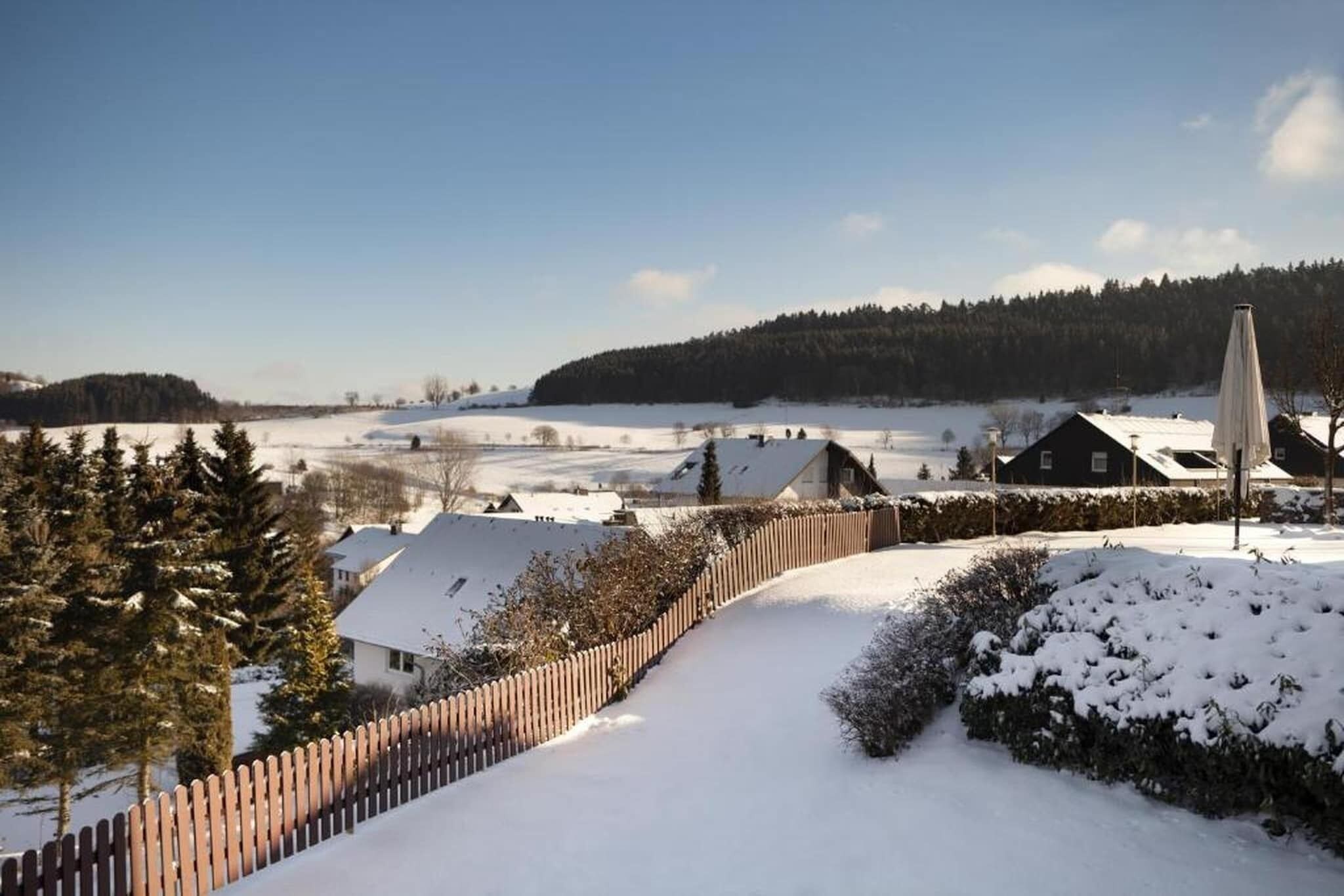 Un paysage enneigé avec une clôture en bois, une maison au toit en pente et une forêt lointaine.