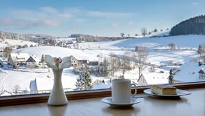 Una mesa con una taza y platillo blancos, un florero blanco con una flor, y una porción de pastel en un plato, con vistas a un paisaje nevado con casas y árboles.
