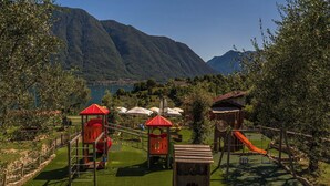 Un parc infantil amb tobogans i elements per escalar, envoltat d'una tanca i arbres.