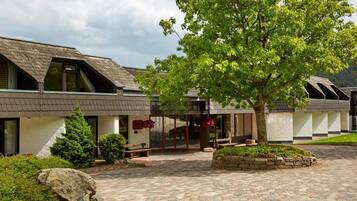 A building with a sloped roof, a covered entrance, and a paved courtyard with a tree and seating area.