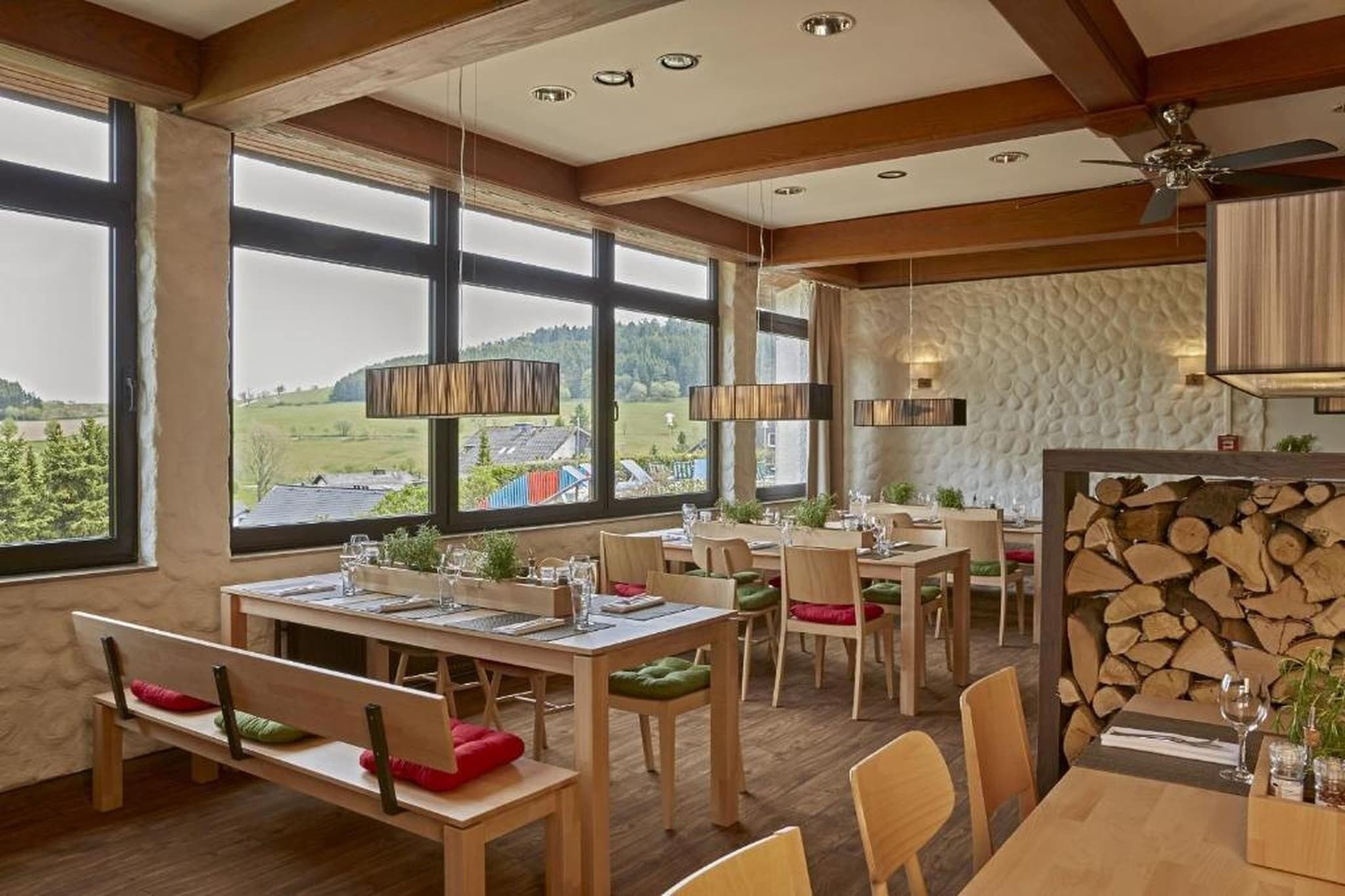 A dining area with wooden tables and chairs, large windows with a view of the countryside, and a ceiling fan.