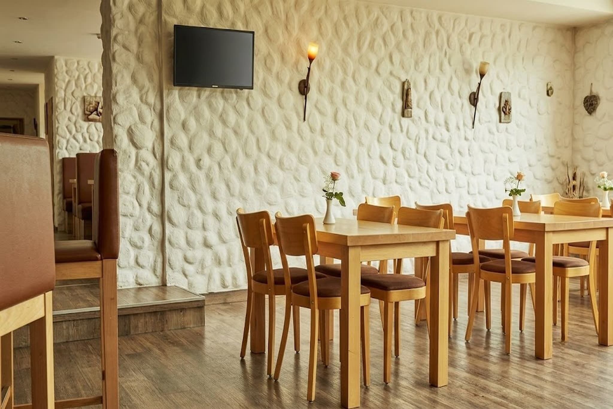 Uma área de jantar com mesas e cadeiras de madeira, uma TV de tela plana na parede e luminárias decorativas.