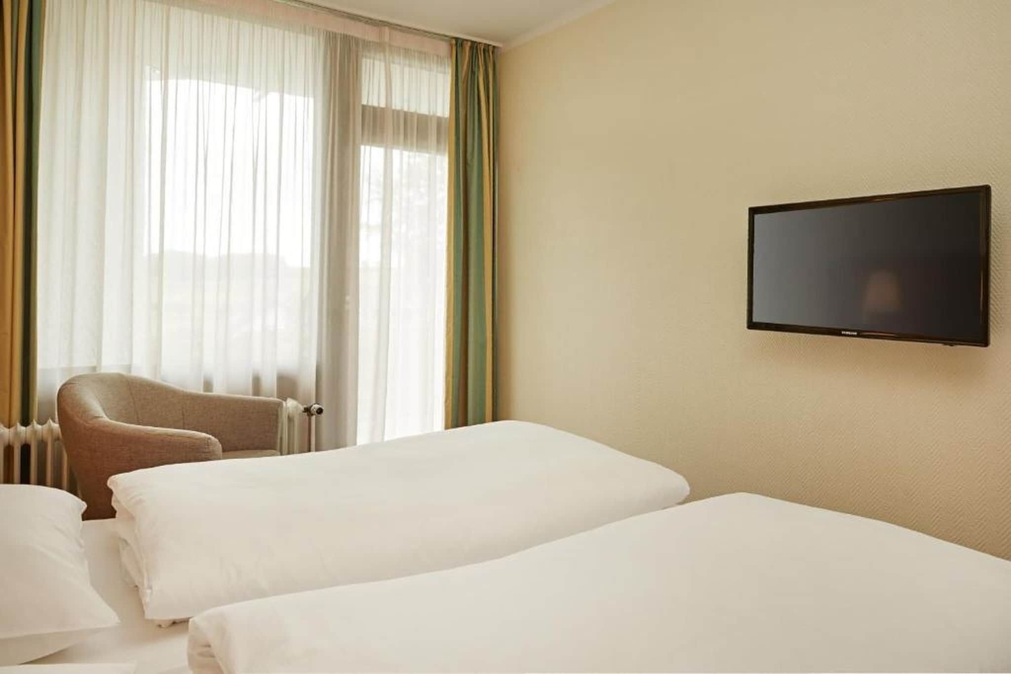 Ein Hotelzimmer mit zwei Betten, einem Wandtelevisor, einem Sessel und einem Fenster mit Vorhängen.