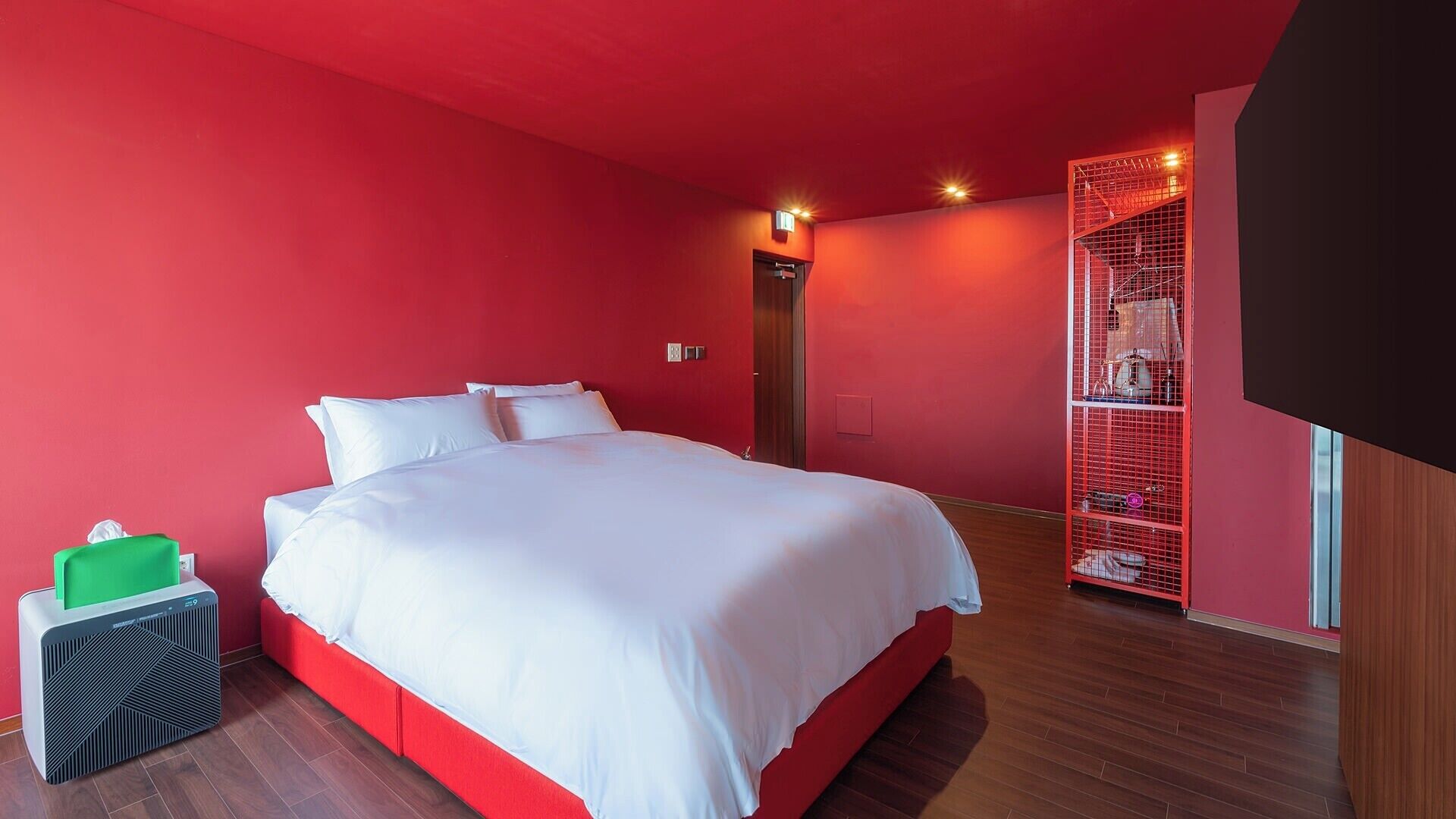 A hotel room with a large bed, a red wall, and a red storage unit.
